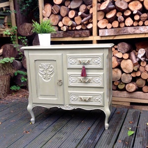 Op de afbeelding: Een vintage, lichtgroene commode met sierlijke houtsnijwerk en een enkele deur aan de linkerkant. De commode heeft drie laden met gouden handvatten en een rode kwast op de bovenste lade. De commode staat op een houten dek.
