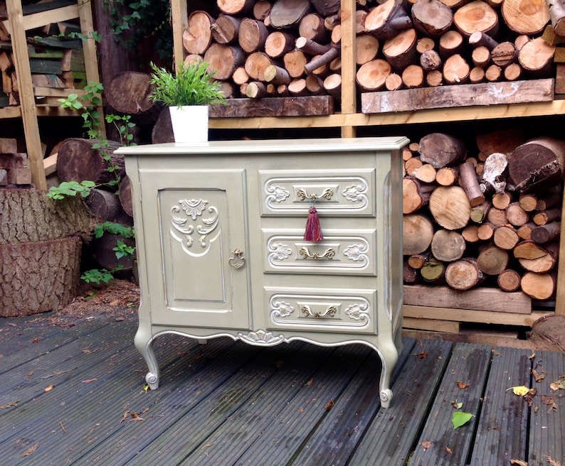 Op de afbeelding: Een vintage, lichtgroene commode met sierlijke houtsnijwerk en een enkele deur aan de linkerkant. De commode heeft drie laden met gouden handvatten en een rode kwast op de bovenste lade. De commode staat op een houten dek.