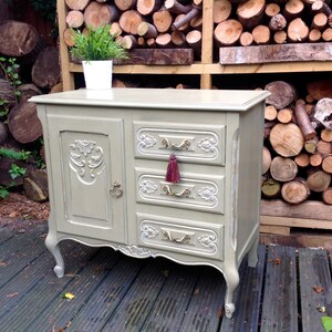 Op de afbeelding: Een vintage-stijl dressoir met een lichtgrijze afwerking en sierlijke details. Het heeft een enkele deur aan de linkerkant en drie lades aan de rechterkant. De lades hebben goudkleurige handvatten en een rode kwast op de middelste lade.