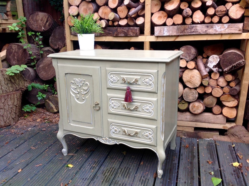 Op de afbeelding: Een vintage-stijl dressoir met een lichtgrijze afwerking en sierlijke details. Het heeft een enkele deur aan de linkerkant en drie lades aan de rechterkant. De lades hebben goudkleurige handvatten en een rode kwast op de middelste lade.