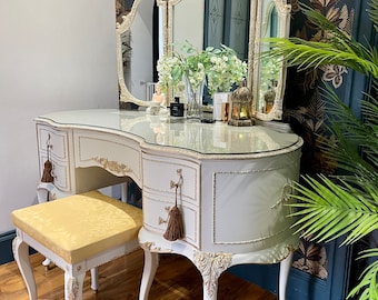 Può includere: Tavolo da toeletta bianco con piano curvo e tre cassetti. Il tavolo ha un piano in vetro e uno specchio a tre pannelli con decorazioni dorate. Uno sgabello basso con una seduta in tessuto giallo si trova davanti al tavolo.
