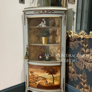 May include: A corner cabinet with a light blue finish and gold accents. The cabinet has a wire mesh door and a painted scene on the bottom panel. The cabinet is decorated with gold trim and has a small tassel hanging from the bottom.