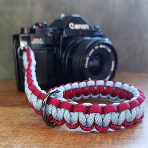 Könnte beinhalten: Ein rotes und graues Paracord-Kameraband mit einem schwarzen Metallring. Das Band ist aus gewebtem Paracord gefertigt und soll strapazierfähig und bequem sein.