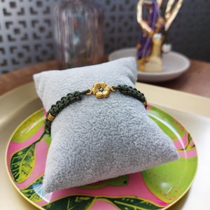 May include: A green braided bracelet with a gold flower charm. The bracelet is displayed on a gray cushion.