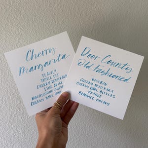 May include: Two square white cards featuring blue handwritten calligraphy. The left card lists Cherry Margarita ingredients: Tequila, Triple Sec, Cherry Demerara, Lime Juice, Maraschino Cherry, and Cherry Bark Vanilla. The right card lists Door County Old Fashioned ingredients: Bourbon, Cherry Demerara, Cherry Bark Bitters, Orange, and Brandied Cherry.