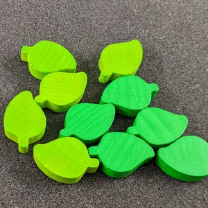 Könnte beinhalten: Zehn grüne, blattförmige Spielsteine aus Holz. Die Steine sind in einem lockeren Kreis angeordnet.