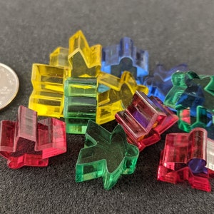 Może przedstawiać: Kolekcja kolorowych, przezroczystych elementów do gry w kolorze czerwonym, żółtym, zielonym i niebieskim. Moneta służy jako odniesienie do rozmiaru. Elementy mają geometryczny, abstrakcyjny kształt i są rozrzucone na ciemnej powierzchni.