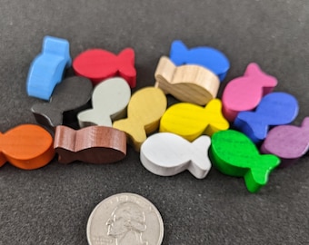 Puede incluir: Una colección de piezas de juego de madera en forma de pez de varios colores, incluyendo naranja, rojo, amarillo, verde, azul, morado, marrón, blanco y negro. Las piezas están dispuestas en una pila sobre una superficie gris oscuro.