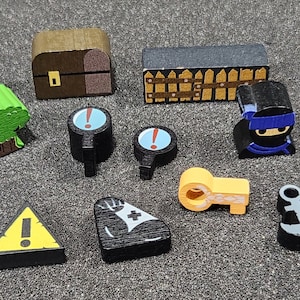 May include: A set of wooden game pieces with various designs, including a tree, a chest, a fence, a key, a ninja, an exclamation point, a muscle, and an anchor. The pieces are painted in different colors, including green, brown, yellow, blue, and black.