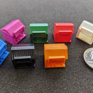 Op de afbeelding: Acht miniatuurgebouwen in verschillende kleuren, waaronder blauw, roze, groen, rood, zwart, wit en oranje. De gebouwen zijn allemaal even groot en in dezelfde vorm, met een plat dak en een klein raam. Ze zijn in een cirkel op een donkergrijs oppervlak gerangschikt.