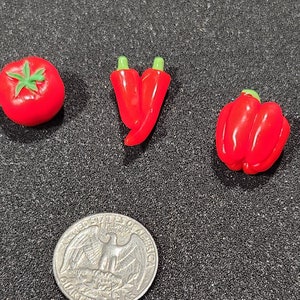 Puede incluir: Tres miniaturas de verduras de plástico rojo: un tomate, un pimiento y dos chiles. Las verduras están sobre una superficie gris oscura.