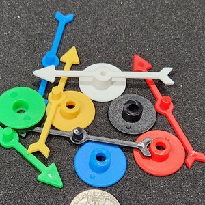 May include: A set of colorful plastic game pieces with arrows and circular bases. The pieces are in various colors, including green, yellow, blue, red, white, and black.