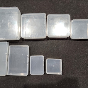 May include: A selection of clear, rectangular plastic storage boxes in different sizes. The boxes are arranged on a dark surface, with lids closed. Sizes vary from large, square boxes to small, square boxes.