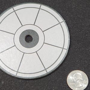 Op de afbeelding: Een wit en grijs cirkelvormig doel met een zwarte middelcirkel en acht spaken. Het doel is ongeveer zo groot als een Nederlandse euro.