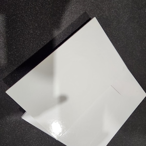 Puede incluir: Dos tableros para tartas blancos apilados uno encima del otro. Los tableros son cuadrados y tienen una superficie lisa y brillante.