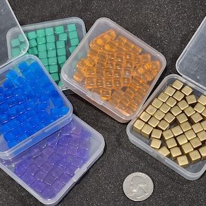 Op de afbeelding: Vijf doorzichtige plastic containers gevuld met kleine, vierkante kralen in verschillende kleuren. De containers zijn in een halve cirkel op een zwart oppervlak gerangschikt. De kralen zijn blauw, groen, oranje, paars en goud.
