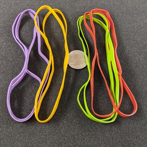 Könnte beinhalten: Vier Gummibänder in verschiedenen Farben: lila, gelb, grün und rot. Die Gummibänder liegen lose auf einer schwarzen Oberfläche. Eine silberne Münze befindet sich in der Mitte des Bildes.