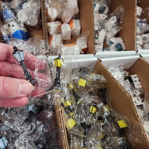 May include: Miniature street lamps with black posts and yellow or light blue lantern tops, individually wrapped in clear plastic packaging. These small decorative items are organized in cardboard bins alongside miniature house figurines.