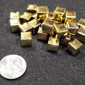 May include: A pile of small, shiny, gold-coloured square beads. A silver coin is next to the beads for size comparison. The beads are scattered on a dark, textured surface.