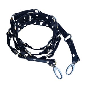 K&ouml;nnte beinhalten: Schwarzer Leder-Taschenriemen mit silberfarbenen Metallringen und -verschl&uuml;ssen. Der Riemen hat ein geflochtenes Design mit kreisf&ouml;rmigen Aussparungen und ist f&uuml;r eine Handtasche konzipiert.