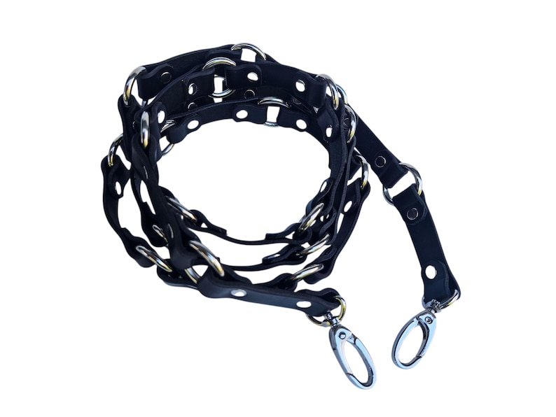 K&ouml;nnte beinhalten: Schwarzer Leder-Taschenriemen mit silberfarbenen Metallringen und -verschl&uuml;ssen. Der Riemen hat ein geflochtenes Design mit kreisf&ouml;rmigen Aussparungen und ist f&uuml;r eine Handtasche konzipiert.