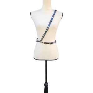 K&ouml;nnte beinhalten: Ein schwarzer Leder-Harness mit silberfarbenen Metallbeschl&auml;gen wird auf einer Schaufensterpuppe pr&auml;sentiert. Der Harness hat einen Schultergurt und einen Tailleng&uuml;rtel, die durch verstellbare Riemen und Metallverschl&uuml;sse verbunden sind. Die Schaufensterpuppe steht auf einem schwarzen St&auml;nder.