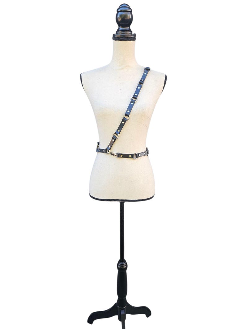 K&ouml;nnte beinhalten: Ein schwarzer Leder-Harness mit silberfarbenen Metallbeschl&auml;gen wird auf einer Schaufensterpuppe pr&auml;sentiert. Der Harness hat einen Schultergurt und einen Tailleng&uuml;rtel, die durch verstellbare Riemen und Metallverschl&uuml;sse verbunden sind. Die Schaufensterpuppe steht auf einem schwarzen St&auml;nder.