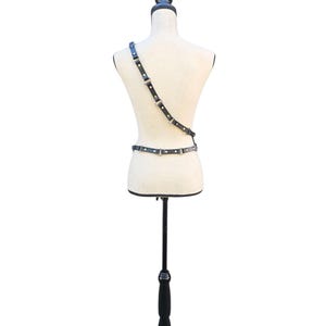 K&ouml;nnte beinhalten: Schwarzer Leder-Harness mit silberfarbenen Beschl&auml;gen, auf einer Schaufensterpuppe pr&auml;sentiert. Der Harness hat einen Schultergurt und einen Tailleng&uuml;rtel, die durch verstellbare Riemen verbunden sind. Die Schaufensterpuppe steht auf einem schwarzen St&auml;nder vor wei&szlig;em Hintergrund.