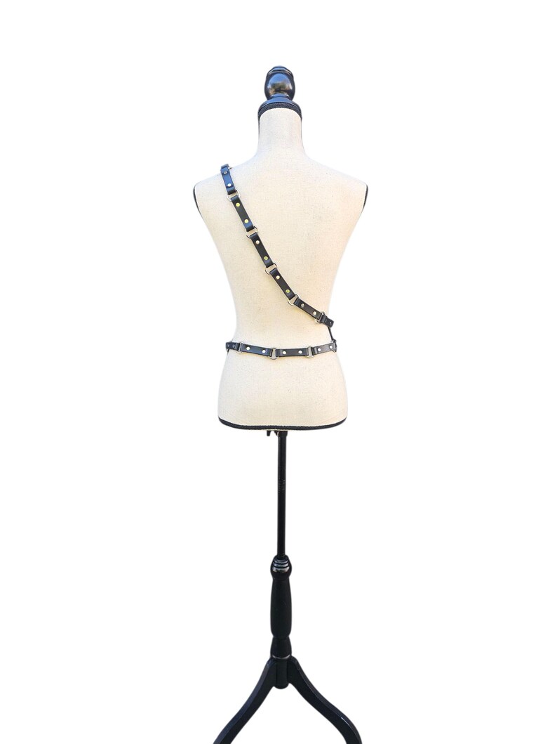 K&ouml;nnte beinhalten: Schwarzer Leder-Harness mit silberfarbenen Beschl&auml;gen, auf einer Schaufensterpuppe pr&auml;sentiert. Der Harness hat einen Schultergurt und einen Tailleng&uuml;rtel, die durch verstellbare Riemen verbunden sind. Die Schaufensterpuppe steht auf einem schwarzen St&auml;nder vor wei&szlig;em Hintergrund.