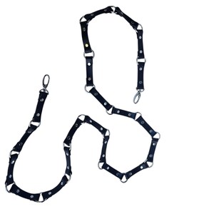 K&ouml;nnte beinhalten: Schwarzes Leder-K&ouml;rpergeschirr mit silberfarbenen Metallringen und Verschl&uuml;ssen. Das Geschirr besteht aus mehreren verbundenen Segmenten, die ein geometrisches Design bilden. Das Design wird vor wei&szlig;em Hintergrund angezeigt.