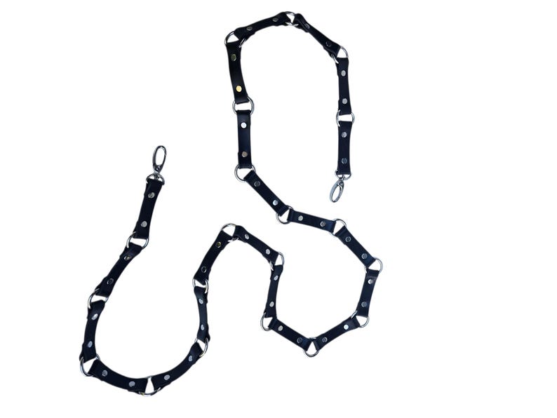 K&ouml;nnte beinhalten: Schwarzes Leder-K&ouml;rpergeschirr mit silberfarbenen Metallringen und Verschl&uuml;ssen. Das Geschirr besteht aus mehreren verbundenen Segmenten, die ein geometrisches Design bilden. Das Design wird vor wei&szlig;em Hintergrund angezeigt.