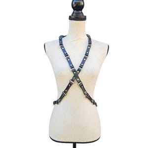 K&ouml;nnte beinhalten: Schwarzes Leder-K&ouml;rpergeschirr mit verstellbaren Riemen und Metallbeschl&auml;gen. Das Geschirr wird auf einer Schaufensterpuppe vor wei&szlig;em Hintergrund pr&auml;sentiert. Die Riemen kreuzen sich vorne und hinten und bilden ein X.