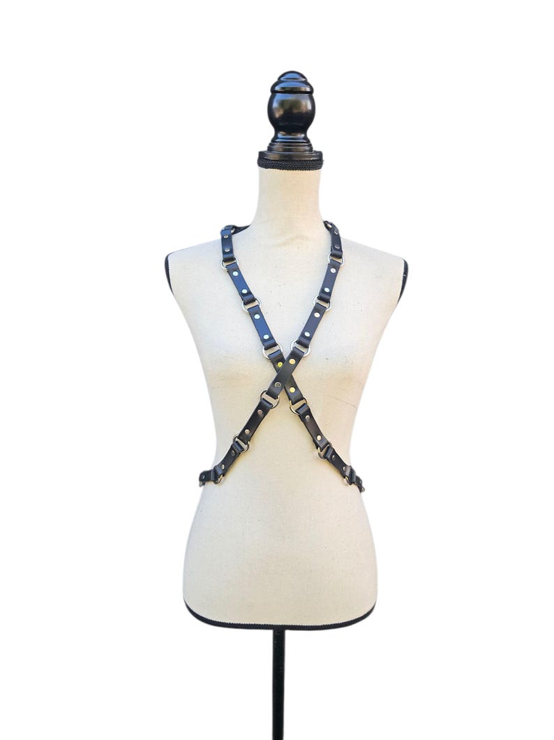 K&ouml;nnte beinhalten: Schwarzes Leder-K&ouml;rpergeschirr mit verstellbaren Riemen und Metallbeschl&auml;gen. Das Geschirr wird auf einer Schaufensterpuppe vor wei&szlig;em Hintergrund pr&auml;sentiert. Die Riemen kreuzen sich vorne und hinten und bilden ein X.