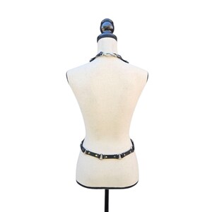 K&ouml;nnte beinhalten: Schwarzes Ledergeschirr mit silberfarbenen Metallbeschl&auml;gen, pr&auml;sentiert auf einer Schaufensterpuppe. Das Geschirr umfasst ein Halsband und einen Tailleng&uuml;rtel. Die Schaufensterpuppe steht auf einem schwarzen St&auml;nder vor wei&szlig;em Hintergrund.