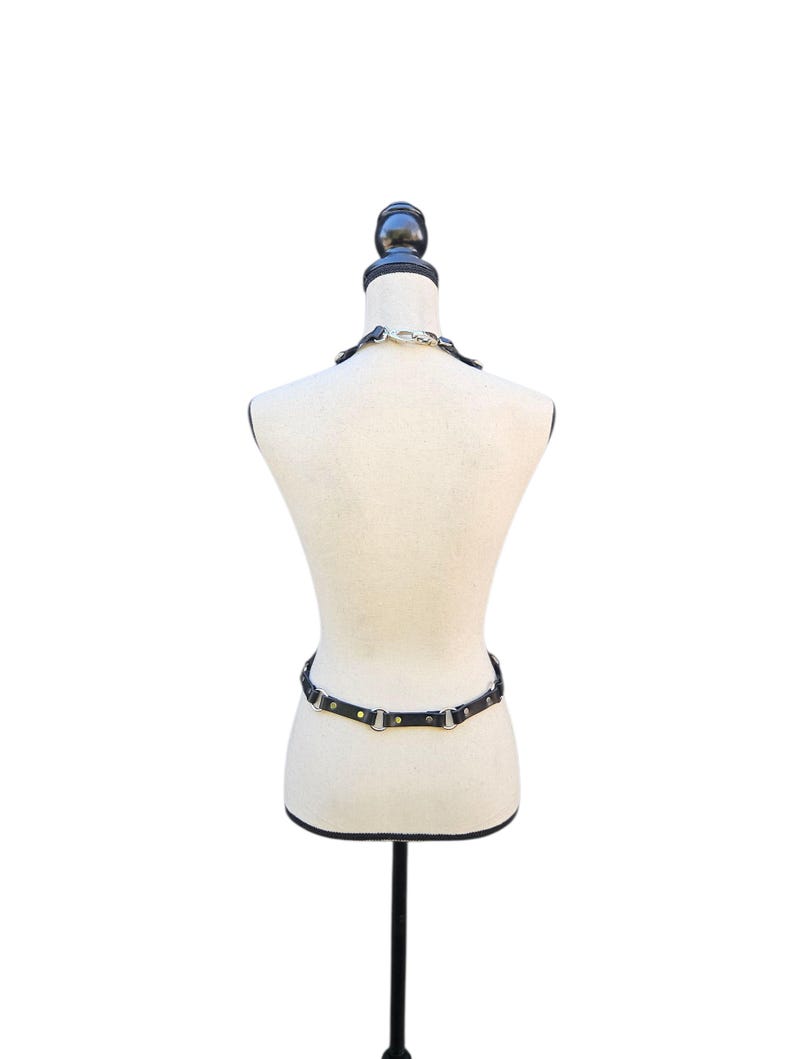 K&ouml;nnte beinhalten: Schwarzes Ledergeschirr mit silberfarbenen Metallbeschl&auml;gen, pr&auml;sentiert auf einer Schaufensterpuppe. Das Geschirr umfasst ein Halsband und einen Tailleng&uuml;rtel. Die Schaufensterpuppe steht auf einem schwarzen St&auml;nder vor wei&szlig;em Hintergrund.