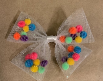 May include: A white tulle bow with colorful pom-poms. The bow is made of a sheer, sparkly fabric and has a white ribbon in the center. The pom-poms are in various colors, including pink, blue, yellow, green, and purple.