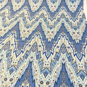May include: A close-up of a fabric with a repeating zigzag pattern in shades of blue, cream, and touches of black. The design incorporates floral and paisley elements, creating a textured, decorative appearance. The fabric is likely used for apparel or home decor.