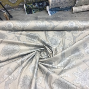 Puede incluir: Un rollo de tela beige con un patr&oacute;n floral plateado. La tela est&aacute; doblada y enrollada, mostrando el patr&oacute;n.
