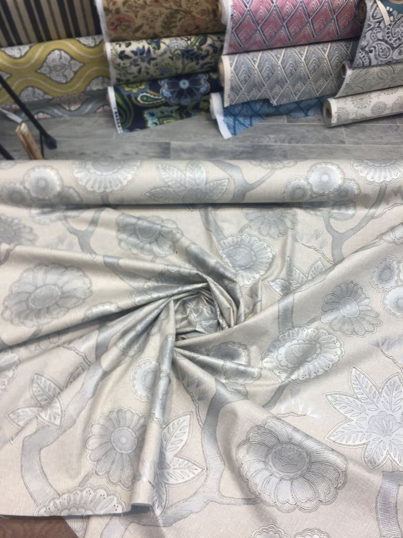 Puede incluir: Un rollo de tela beige con un patr&oacute;n floral plateado. La tela est&aacute; doblada y enrollada, mostrando el patr&oacute;n.