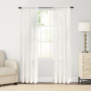 May include: A pair of white sheer curtains hang in a window, with a white wall and a white couch in the background.