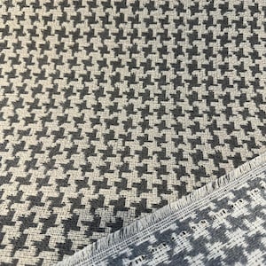 May include: Close-up of a houndstooth fabric in shades of gray and off-white. The textile features a classic pattern of interlocking shapes, with a visible woven texture. The edge of the fabric is frayed, revealing the weave.
