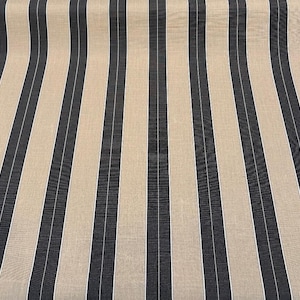 May include: Fabric with a repeating vertical stripe pattern. Alternating stripes of beige, navy blue, and thin white lines create a textured effect. The fabric is slightly curved, suggesting it is draped.