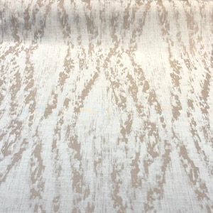 May include: A roll of fabric with a cream-colored background and a pattern of tan, brushstroke-like markings. The fabric is folded over, showing the texture and design. The pattern has a vertical orientation.