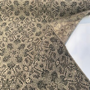 May include: A roll of fabric featuring a repeating damask pattern in black and beige. The textile displays intricate floral and scrollwork motifs across the surface, with a folded corner revealing the reverse side of the woven material.