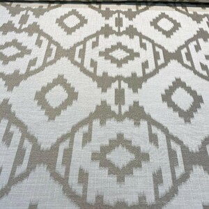 May include: Close-up of a fabric roll with a geometric pattern. The design features diamond and rectangular shapes in a neutral color palette of beige and white. The fabric has a textured appearance, suitable for upholstery or home decor projects.