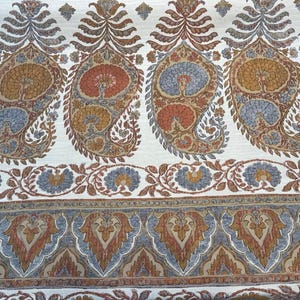 May include: Close-up of a textile with a repeating paisley pattern in shades of rust, gold, and blue on a cream background. The design includes floral and leaf motifs, creating a detailed and ornate look. The fabric has a decorative border.
