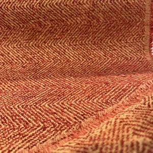 Puede incluir: Primer plano de una tela texturizada con un patrón de espiga. La tela presenta una mezcla de colores rojo y crema, creando una estética cálida. Los bordes de la tela están deshilachados, añadiendo un toque rústico. Este textil es adecuado para la decoración del hogar.