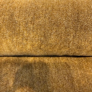 Puede incluir: Una vista de cerca de una pieza de tela doblada con una superficie densa, difusa y texturizada. El material es de un color marrón dorado cálido y sólido con variaciones sutiles de tono en la suave fibra del tejido.