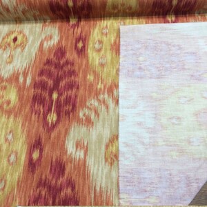 May include: Close-up of a fabric swatch with an orange, red, and beige ikat pattern. The fabric is shown with the back side facing up, revealing a white, textured backing.