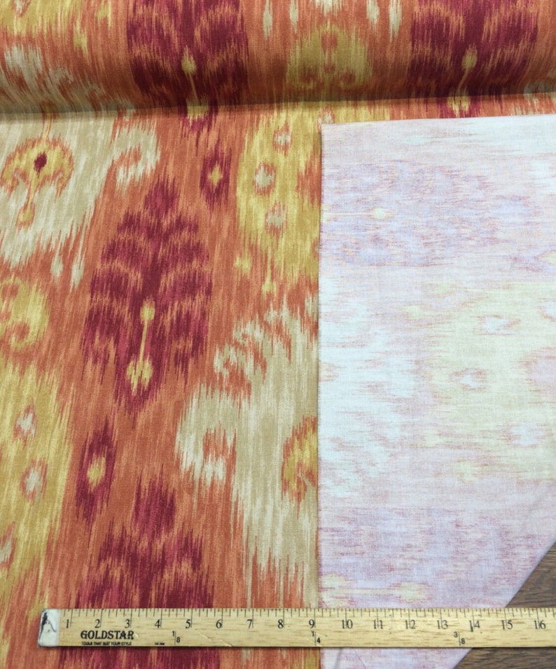 May include: Close-up of a fabric swatch with an orange, red, and beige ikat pattern. The fabric is shown with the back side facing up, revealing a white, textured backing.